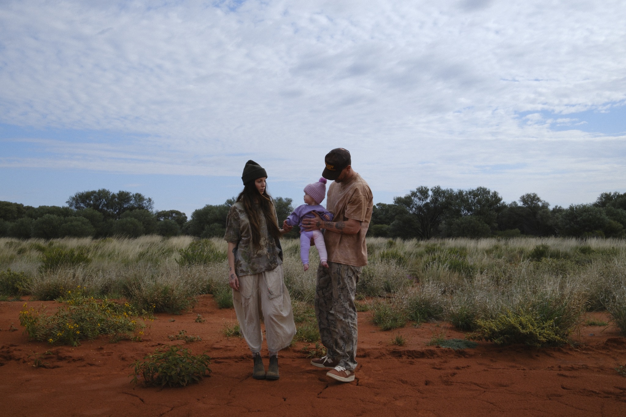 Family of three together on red earth among desert grasses beneath a cloudy sky